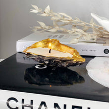 Load image into Gallery viewer, Hand poured shell candle made from recycled oyster shell with gold tip, resting on a stylish book, illuminating its surroundings.